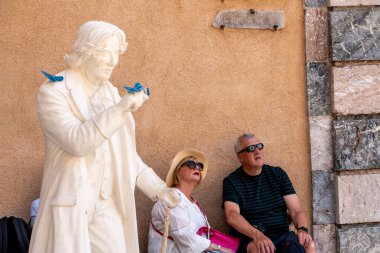 Taormina, Sizilien - İtalya - 05-04-2025: Mavi kelebekli Oscar Wilde heykeli ve Sicilya 'nın Taormina kentindeki dokulu bir duvarın yanında mermer bir taş sütunun yanında oturan iki turist.
