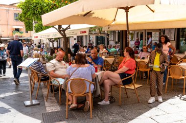 Taormina, Sizilien - İtalya - 05-04-2025: Sicilya 'nın Taormina kentindeki açık hava restoran sahnesi, şemsiyelerin altında oturan misafirler, yürüyen insanlar ve masaların yanına su döken bir çocuk.
