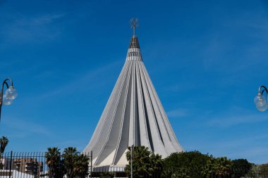 Syrakus, Sizilien - İtalya - 04-09-2025: Syracuse 'daki uzun konik bazilika Madonna delle Lacrime tırtıklı beton yapısı, merkezi kulesi, çiti, palmiye ağaçları ve mavi gökyüzü altında sokak lambaları.