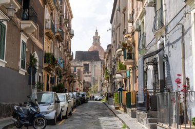 Catania, Sizilien - İtalya - 04-15-2025: Catania 'da dar sokaklar, park edilmiş araçlar ve eski binalar, uzak mesafede büyük bir kubbe bulunan bir kilisede son buluyor..