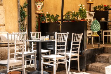 Taormina, Sizilien - İtalya - 05-04-2025: Taormina, Sicilya 'da açık kafe, beyaz sandalyeler, siyah masa, bardaklar, çatal bıçak, seramik süslemeler ve parlak güneş ışığı altında çiçekler.