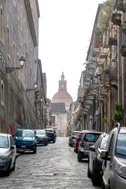 Catania, Sizilien - İtalya - 04-15-2025: Dar kaldırım taşı caddesi arabalarla ve tarihi binalarla kaplı, uzak arkaplanda görünen büyük bir kubbe yapısıyla sona eriyor.