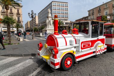Catania, Sizilien - İtalya - 04-15-2025: Kırmızı ve beyaz bir turist treni, Catania, Sicilya 'da palmiye ağaçları, yayalar ve kentsel binaların bulunduğu Vincenzo Bellini anıtının önünden geçiyor.