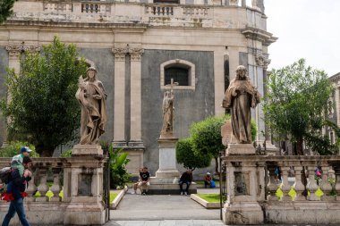 Catania, Sizilien - İtalya - 04-15-2025: Catania 'da bir kilise duvarının önündeki üç taş heykel, ağaçlar, banklarda oturan ziyaretçiler ve arka planda mimari unsurlar.