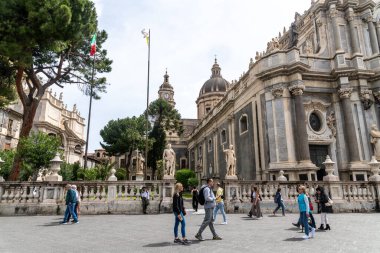 Catania, Sizilien - İtalya - 04-15-2025: İnsanlar Catania 'daki tarihi katedralin önünden geçerek, süslü heykeller, kubbe mimarisi, ağaçlar, sütunlar ve dalgalanan ulusal bayrakların yer aldığı.