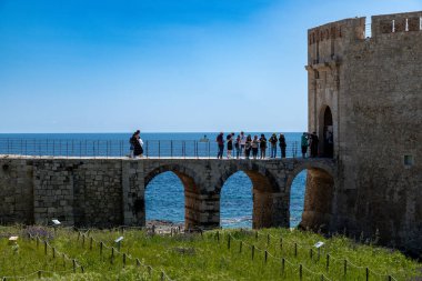 Syrakus, Sizilien - İtalya - 04-09-2025: Güneşli Siraküza 'da bir kıyı kalesine bağlı tarihi bir taş köprüde yürüyen ve ayakta duran bir grup turist.