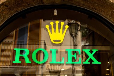 Taormina, Sizilien - İtalya - 05-04-2025: Taormina, Sicilya 'daki Rolex mağazasının penceresine yakın plan, binalar ve lamba yansımaları içeren yeşil Rolex metin ve sarı taç logosunu gösteriyor.
