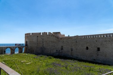 Syrakus, Sizilien - İtalya - 04-09-2025: Siraküza 'daki tarihi taş kale kemerli köprü, çimenli hendek ve açık mavi gökyüzü altında okyanus manzaralı, bir grup turist mevcut.