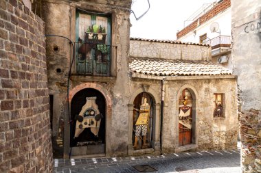 Graniti, Sizilien - İtalya - 05-05-2025: Boyalı ahşap figürler, Graniti, Sicilya 'da güneş ışığıyla aydınlatılan taştan bir evin kemerli kapılarını ve balkonunu süslüyor.