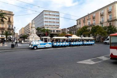Catania, Sizilien - İtalya - 04-15-2025: Mavi ve beyaz turist treni, Catania şehir merkezinde heykel, modern ve tarihi binalar ve yayaların bulunduğu işlek bir şehir meydanından geçiyor.