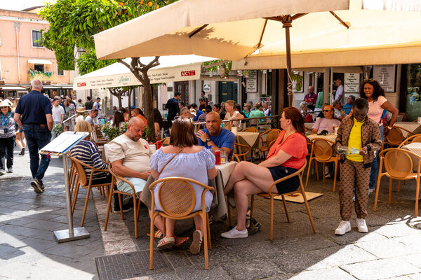 Таормина, Сицилия - Италия - 05-04-2025: сцена ресторана на открытом воздухе в Таормине, Сицилия с сидячими гостями под пумбреллами, людьми, проходящими мимо, и ребенком, льющим воду возле столов.