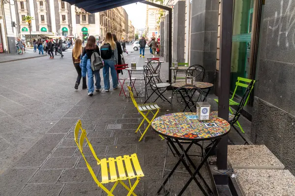 Catania, Sizilien - İtalya - 04-15-2025: Renkli fayansları ve katlanabilir sandalyeleri olan açık hava kafe masaları yayalar Catania, Sicilya 'da işlek bir şehir caddesinde yürürken kaldırımda sıralanır.