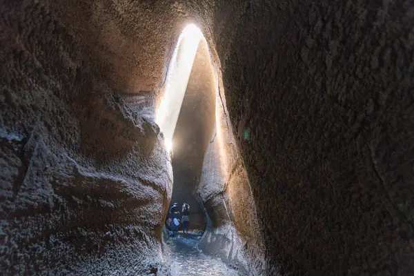 Linguaglossa, Sizilien - İtalya - 05-01-2025: Dar bir volkanik mağaranın içinde, insanlar karanlık geçidi izlerken, güneş ışığı tavan yarığından içeri girer ve pürüzlü duvarları aydınlatır..