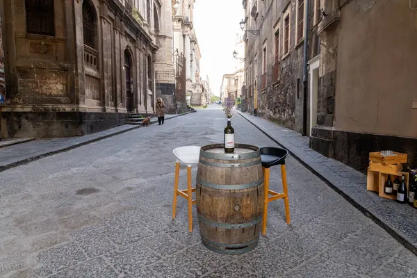 Catania, Sizilien - İtalya - 04-15-2025: Dar Catania caddesinde sandalyeler ve şarap şişesi bulunan şarap fıçısı masası, yaşlanmış binalar ve güneş ışığında yürüyen köpek tarafından çevrelenmiş.