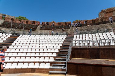Taormina, Sizilien - İtalya - 05-04-2025: Taormina, Sicilya 'daki antik tiyatro manzarası, modern beyaz koltuklar, eski tuğla kalıntıları ve açık mavi gökyüzünün altında yürüyen ziyaretçileri gösteriyor.