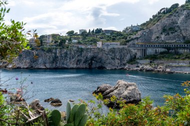 Taormina, Sizilien - İtalya - 05-04-2025: Sahil kayalıkları, sahil binaları ve Taormina yakınlarındaki sakin deniz manzarası, görünür bitki örtüsü ve kayalık ön plan ayrıntılarıyla.