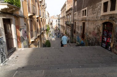 Catania, Sizilien - İtalya - 04-15-2025: Catania, Sicilya 'da duvar yazısıyla kaplı binalar, taş merdivenler, dar yollar, balkonlar, park edilmiş arabalar ve gün ışığında yayalar.