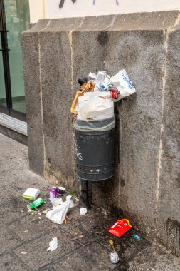 Catania, Sizilien - İtalya - 04-15-2025: Üssünün etrafında yiyecek ambalajı ve dağınık çöp dolu bir çöp kutusu Sicilya 'nın orta kesimindeki taş bir duvara karşı duruyor.