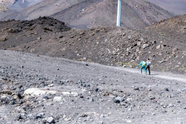 Catania, Sizilien - İtalya - 04-22-2025: Sırt çantaları ve yürüyüş direkleriyle iki yürüyüşçü, Etna Dağı 'nda kayalık arazi ve karanlık yamaçlarla çevrili engebeli bir volkanik patika boyunca yürüyor.