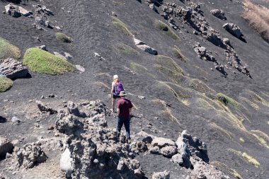 Linguaglossa, Sizilien - İtalya - 05-01-2025: Dağılmış kayalar ve parlak gün ışığında küçük yeşil yosun parçalarıyla siyah bir volkanik yamaçta iki yürüyüşçü yokuş yukarı yürüyor.