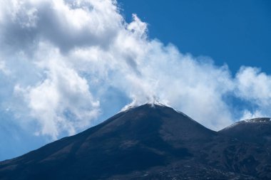 Etnas Dağı 'nın karanlık zirvesi, Sicilya' da bulutlar ve engebeli volkanik arazilerle çevrili mavi gökyüzüne yoğun bir duman bulutu yayar..