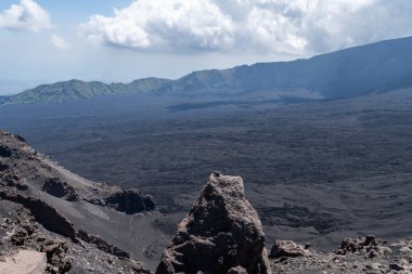 Sicilya 'daki Etna Dağı' nın kayalık krater yamaçlarından uzanan geniş karanlık volkanik düzlükler yeşil tepeler ve uzak mesafelerde bulut örtüsüyle kaplıdır..