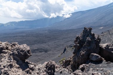 Etna Dağı 'ndaki volkanik plato koyu lav arazisiyle ön plandaki kaya oluşumlarına ve Sicilya' daki dağlık yamaçlardaki bulutlara çarpıyor..