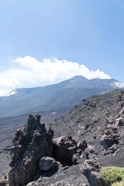 Volkanik yamaç, lava kayaları ve mavi gökyüzünün altında seyrek bitki örtüsüyle dağılmış durumda. Zirve uzak arkaplanda duman yayıyor..