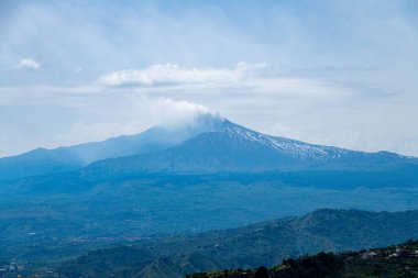 Sicilya, Taormina 'dan Etna Dağı' nın manzarası. Orman ve tepelerin üzerindeki bulutlu gökyüzünün altında kraterden yükselen dumanla birlikte..