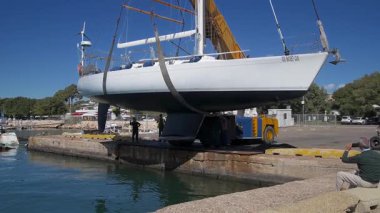 Cagliari - Sardinien - Italien - 03-10-2025: Beyaz bir yelkenli liman sularının üzerindeki sarı bir vinç tarafından denize kontrollü bir kalkış için hazırlanıyor..