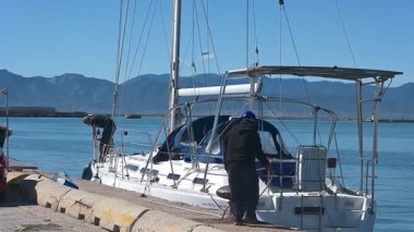 Cagliari - Sardinien - Italien - 03-10-2025: İki adam Peri Masalı adlı beyaz bir yelkenliyi halatlarla, sakin mavi sularla ve arka planda dağlarla limana bağladılar.