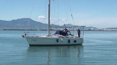 Cagliari - Sardinien - Italien - 03-10-2025: Fairytale adlı beyaz bir yelkenli limana yanaşıyor, güvertede iki mürettebat var, etrafı sakin sular ve bir dağ zeminiyle çevrili..