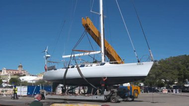 Cagliari - Sardinien - Italien - 03-10-2025: İşçiler, açık mavi gökyüzünün altındaki bir marina şantiyesinde sarı bir vinç tarafından çekilen beyaz yelkenli gibi kaldırma kayışlarına rehberlik ediyorlar.