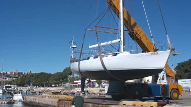 Cagliari - Sardinien - Italien - 03-10-2025: Vinçle askıya alınmış bir yelkenli, açık mavi bir gökyüzünün altında limandan izleyen gözlemcilerle yavaşça limana doğru hareket ediyor..