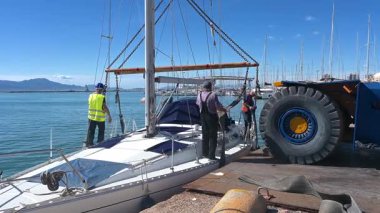 Cagliari - Sardinien - Italien - 03-10-2025: Birkaç işçi mavi gökyüzünün altında liman kenarında vinç kaldırma işlemi hazırlarken demirli bir yata zincir ve teçhizat taşıdılar.