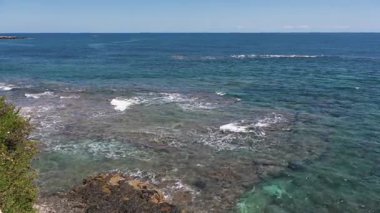 Dalgalar sahil kenarındaki düz kayaların üzerinden süzülerek uzak ufuk çizgisine sahip berrak bir gökyüzünün altında şeffaf turkuaz sularda hafif köpük desenleri oluşturur..