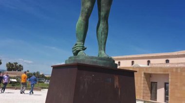 Syrakus, Sizilien - İtalya - 04-09-2025: Pan yukarı doğru Mitoraj heykeli, İkaria grande, Syracuse 'da, mavi gökyüzünün altındaki kaidede bronz kanatlı bir figür ve arka planda binalar ve bitkiler bulunuyor.
