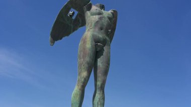 Syrakus, Sizilien - İtalya - 04-09-2025: Pan aşağı doğru Mitoraj heykeli, İkaria grande, Syracuse 'da, mavi gökyüzünün altındaki kaidede bronz kanatlı bir figür ve arkasında binalar ve bitkiler var.