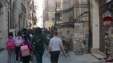 Syrakus, Sizilien - İtalya - 04-08-2025: Bir grup öğrenci Syracuse 'da dar bir sokakta yürüyor, eski cephelerden, açık dükkanlardan ve aksesuar ve hediyelik eşya satan sokak satıcılarından geçiyor.