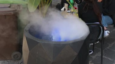 Syrakus, Sizilien - İtalya - 04-08-2025: Bir çokgen siyah baş heykeli, şehir dışındaki bir oturma alanında masa ve bitkilerin yanına yerleştirilmiş, mavi ışıklı yapay sis yayıyor.