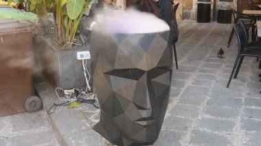 Syrakus, Sizilien - İtalya - 04-08-2025: Bir geometrik siyah baş heykeli, Syracuse 'da bir kaldırım taşı caddesindeki kafeterya tablolarının yanına buhar yayıyor..