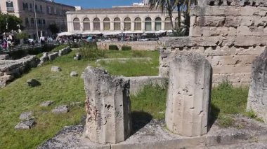 Syrakus, Sizilien - İtalya - 04-08-2025: Syracuse 'deki Apollon tapınağının taş kalıntıları, kırık kolonlar, antik taş duvarlar, çimenli zemin ve arkasında canlı bir sokak pazarı.