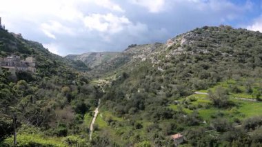Teraslı yamaçları, virajlı yolları, tepeleri ve dramatik bulutlu gökyüzünün altında dağınık kırsal evleri olan bir vadi manzarası..