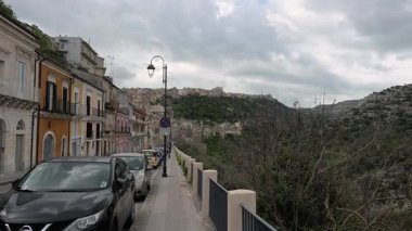 Ragusa, Sizilien - İtalya - 04-03-2025 Redaktionell: Ragusa İbla 'daki tarihi cadde, teraslı yamaçlar ve bulutlu bir dağ vadisine bakan eski binalar ve park edilmiş arabalarla dolu.