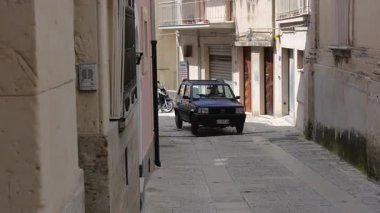 Ragusa, Sizilien - İtalya - 04-03-2025 Redaktionell: Arabalar güneşli bir günde Ragusa 'da dar bir geçitten, eski binalardan, kapılardan ve park etmiş bir scooter' dan geçiyor.