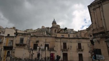 Ragusa, Sizilien - İtalya - 04-03-2025 Redaktionell: Ragusa İbla 'daki tarihi binalar ve kilise kubbesi bulutlu bir gökyüzünün altında, küçük balkonları, kapalı dükkanları ve sessiz bir meydanı gösteriyor