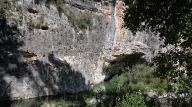 Pantalica 'da yoğun bitki örtüsü, gölgeler ve açık mavi gökyüzü tarafından çerçevelenmiş dar bir derenin üzerinde oyulmuş oyukları olan kayalık uçurumlar yükseliyor..