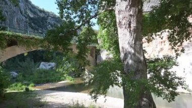 Uçurumlar, yoğun yeşillikler ve çakıl taşlarını çevreleyen gölgeli ağaçlarla dolu sakin bir nehri geçen bir taş kemer köprüsünün panoramik görüntüsü.