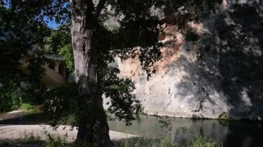 Uçurumlar, yoğun yeşillikler ve çakıl taşlarını çevreleyen gölgeli ağaçlarla dolu sakin bir nehri geçen bir taş kemer köprüsünün panoramik görüntüsü.