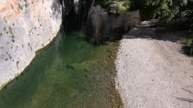 Ağaçlar, gölgeler ve engebeli taş oluşumlarıyla çevrili Pantalica 'da çakıllı kıyıları olan açık yeşil bir dere üzerinde kayalıklar yükseliyor..